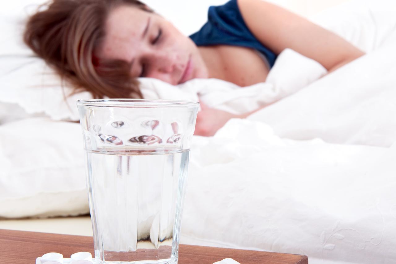 Glass of water and two strips of pills on a bedside table, with a sick woman sleeping in the background